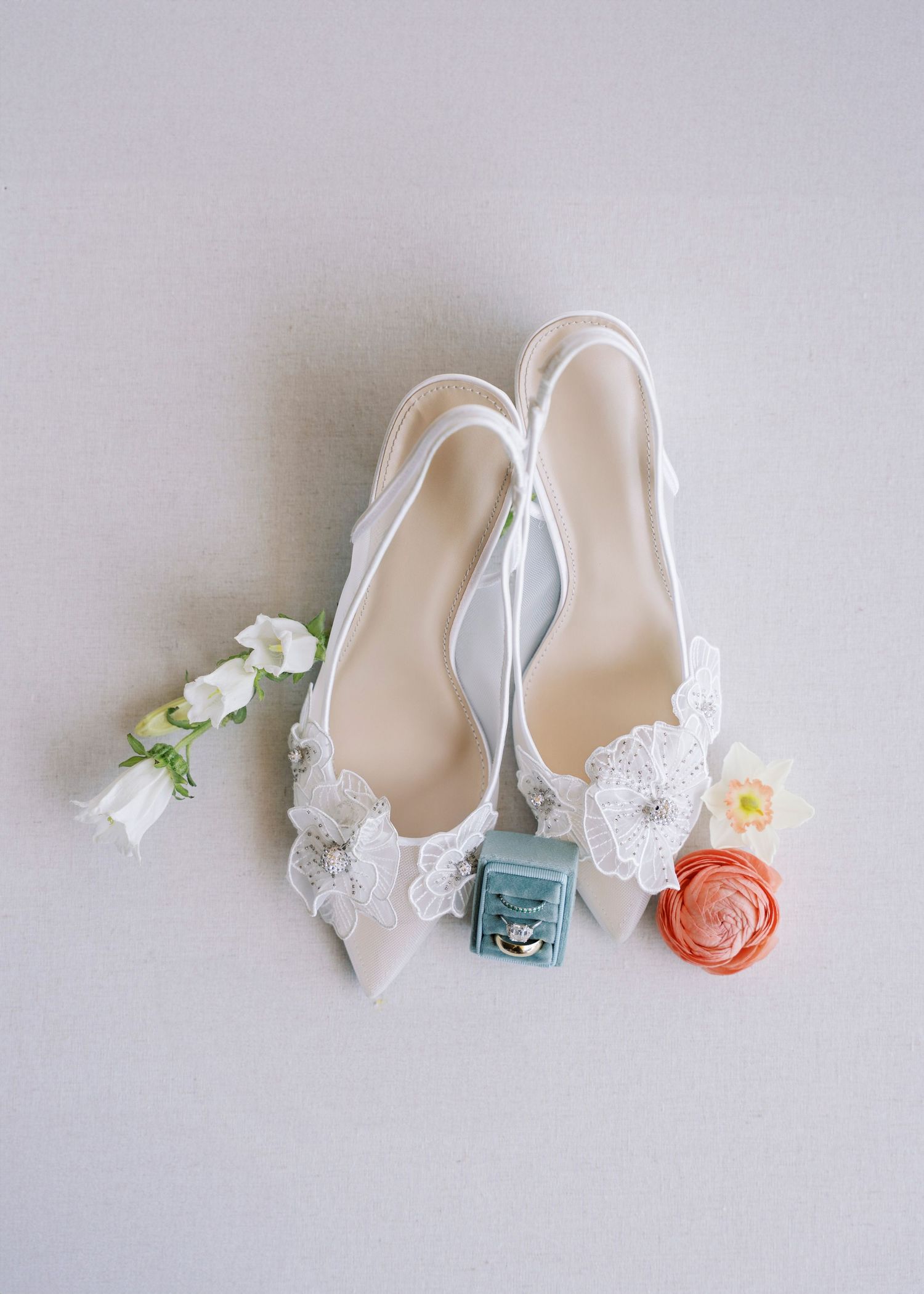 White lace bridal heels displayed with a ring box and colorful flowers on a neutral background.