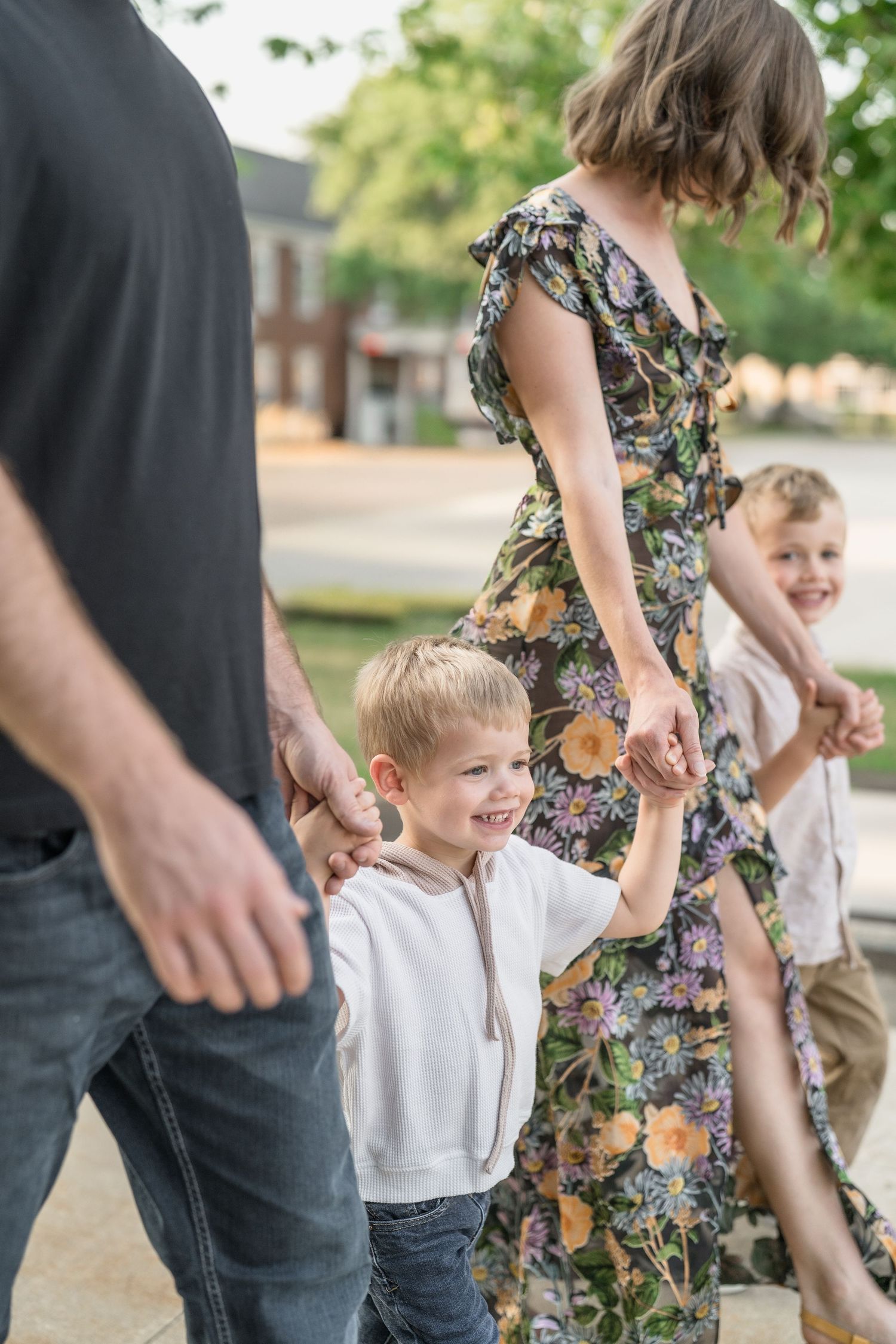 A family holds hands while walking together outdoors, with the woman wearing a floral dress for family photo session in Cuyahoga Falls, Ohio