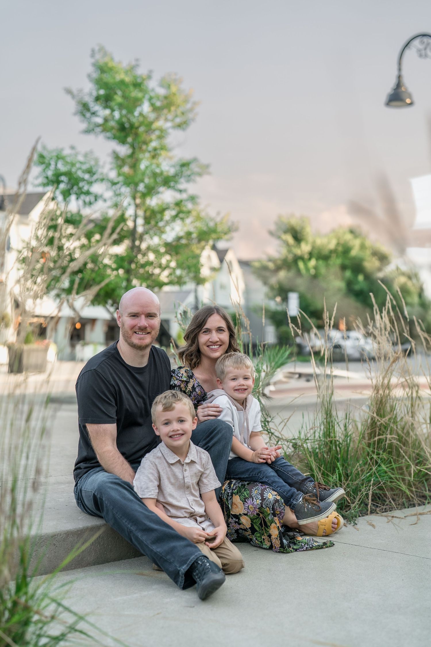 A family sits together outdoors on a curb with trees and shops in the background during golden hour lighting with Cuyahoga Falls, OH Photog