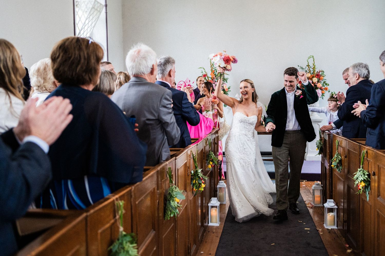 Wedding couple celebrates with guests while walking down church aisle decorated with floral accents during ceremony.