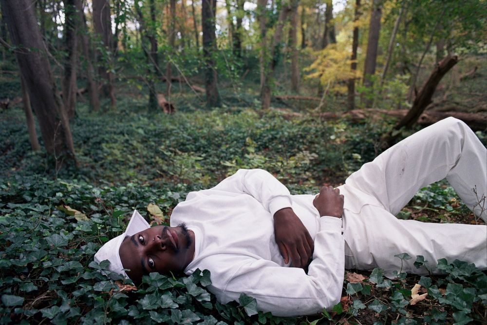 Man in white outfit lying peacefully among green ivy in a lush forest setting.