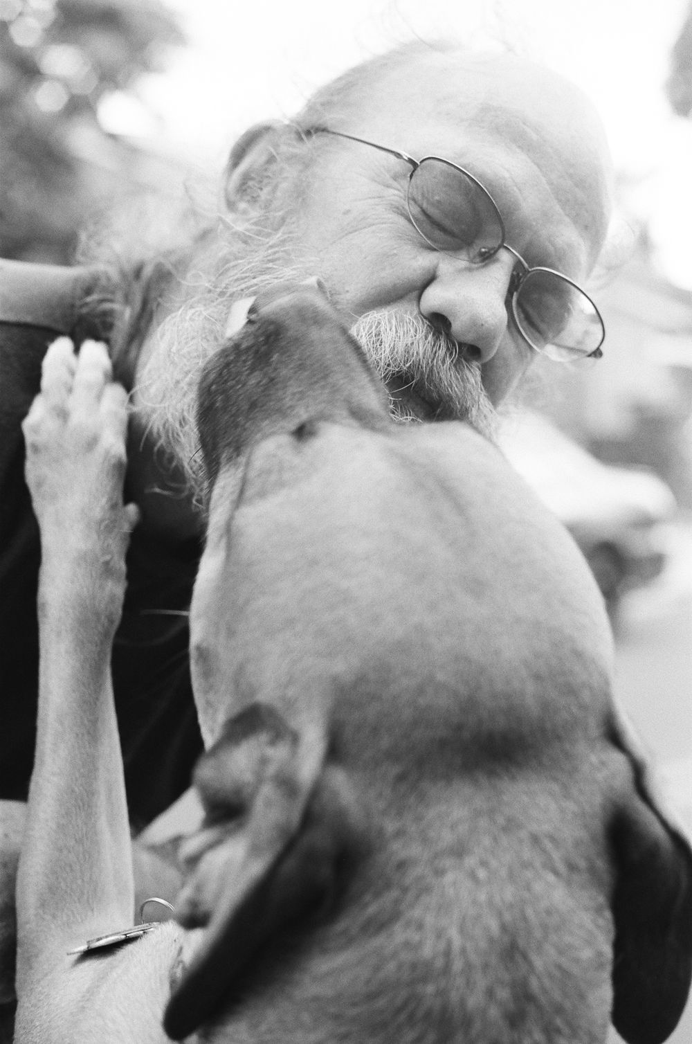 Black and white photo of bearded man with glasses sharing tender moment with dog licking his face outdoors.