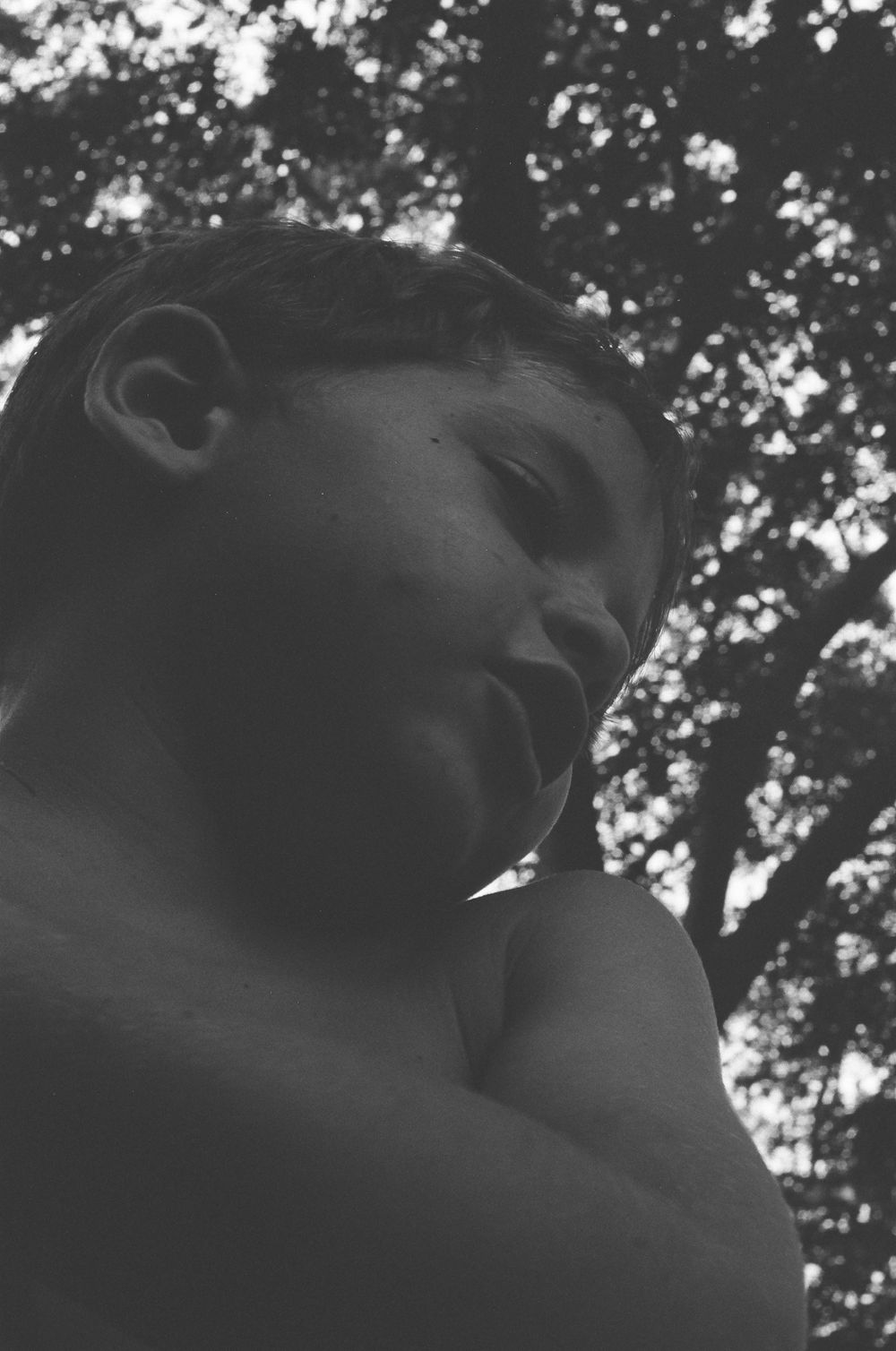 Black and white low-angle portrait of a young boy looking down, with blurred trees in the background.