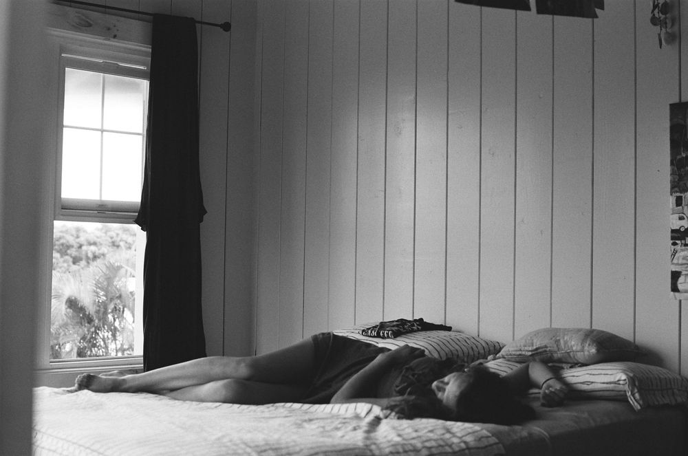 Black and white photo of a person lying on a bed near a window in a rustic white wood-paneled bedroom.