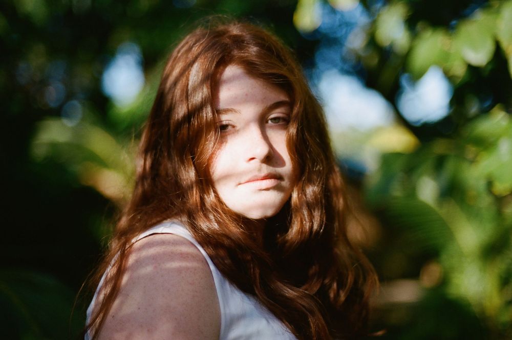Young woman with long auburn hair posing outdoors among green foliage in soft sunlight.
