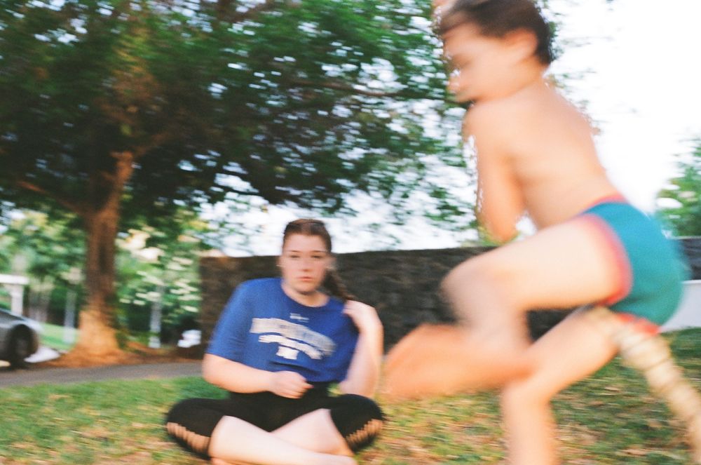 Child jumping energetically outdoors while another child sits cross-legged on grass near trees in blurred motion photo.