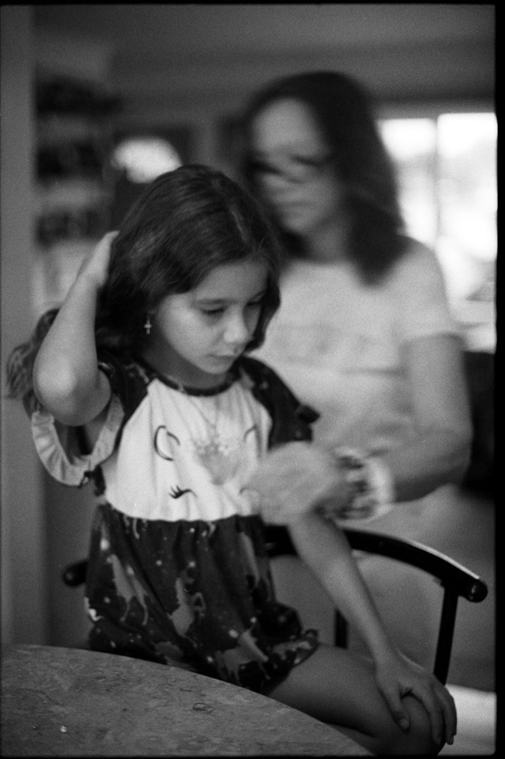 Black and white photo of a young girl having her hair done by a woman in glasses indoors.
