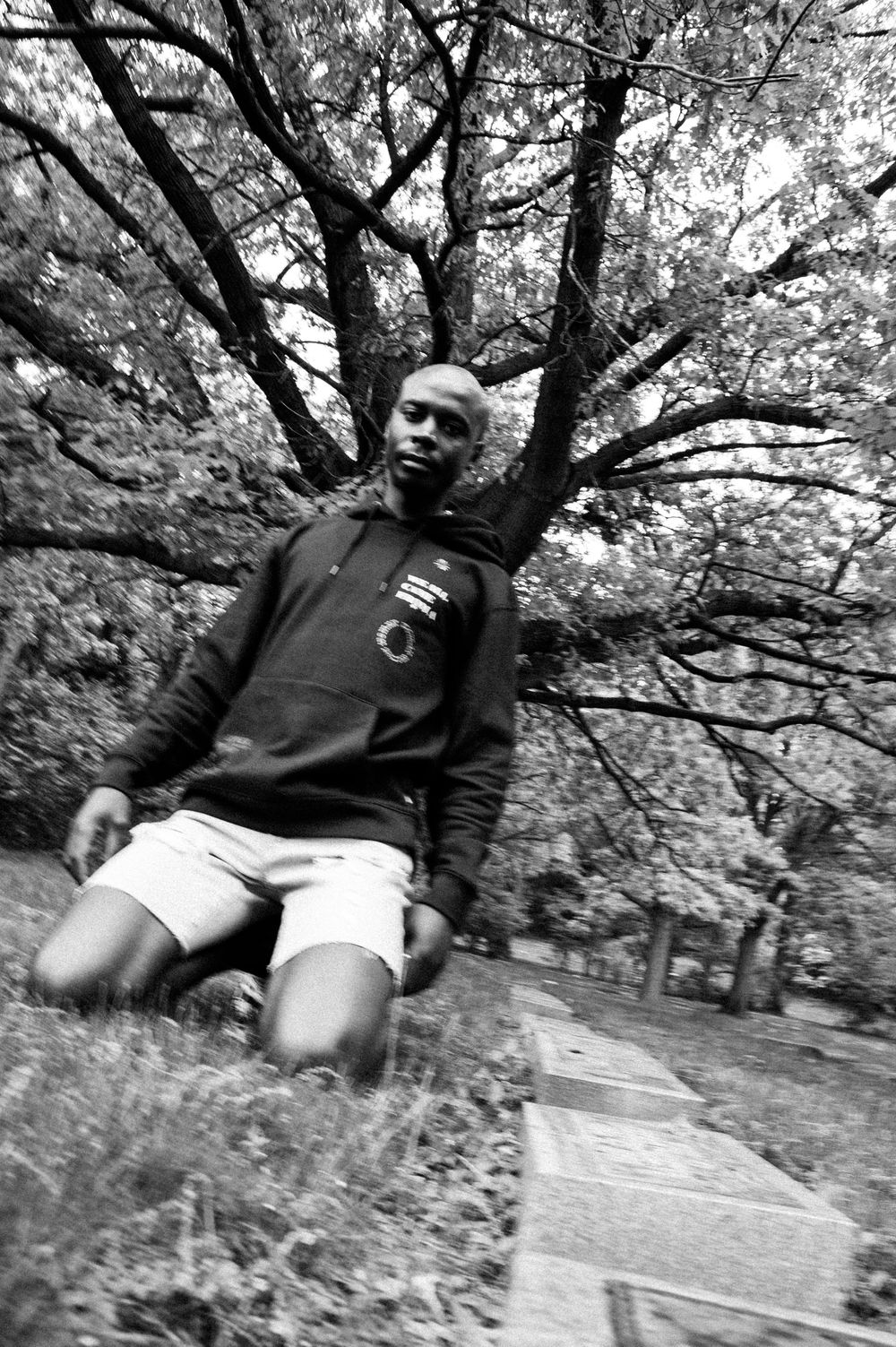 Black and white photo of a man in hoodie and shorts kneeling on grass beneath sprawling trees in a park.