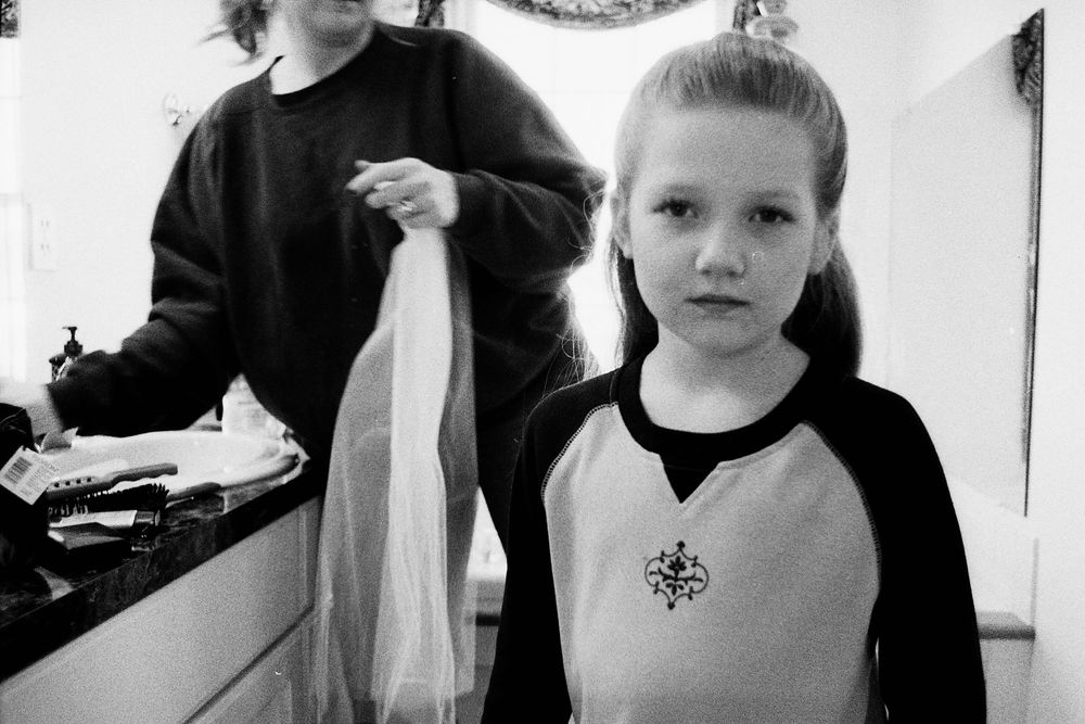Black and white photo of a young girl holding fabric while an adult stands behind her indoors.
