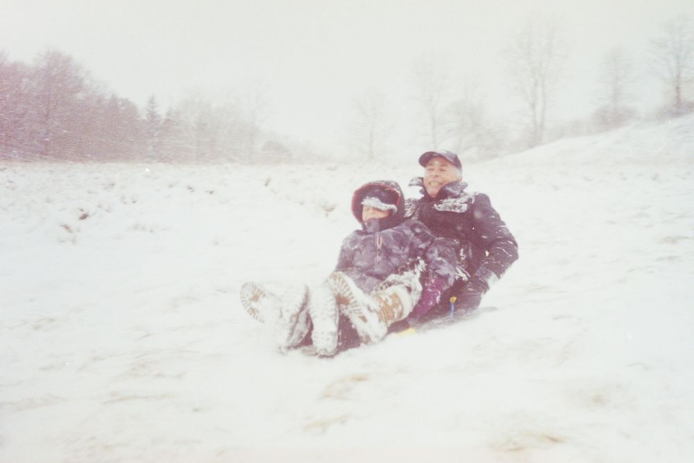 Two people sledding down a snowy hill together during a winter snowstorm in a scenic outdoor landscape.