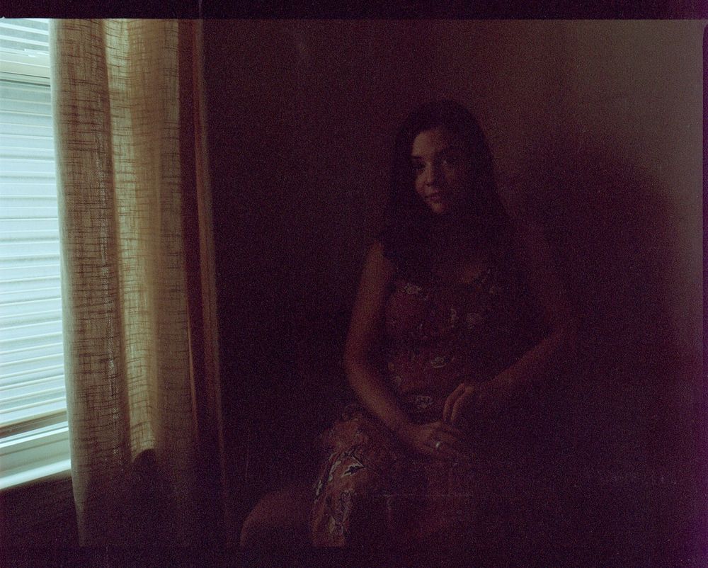A woman with long dark hair sits in a dimly lit room wearing a sequined dress near a window.
