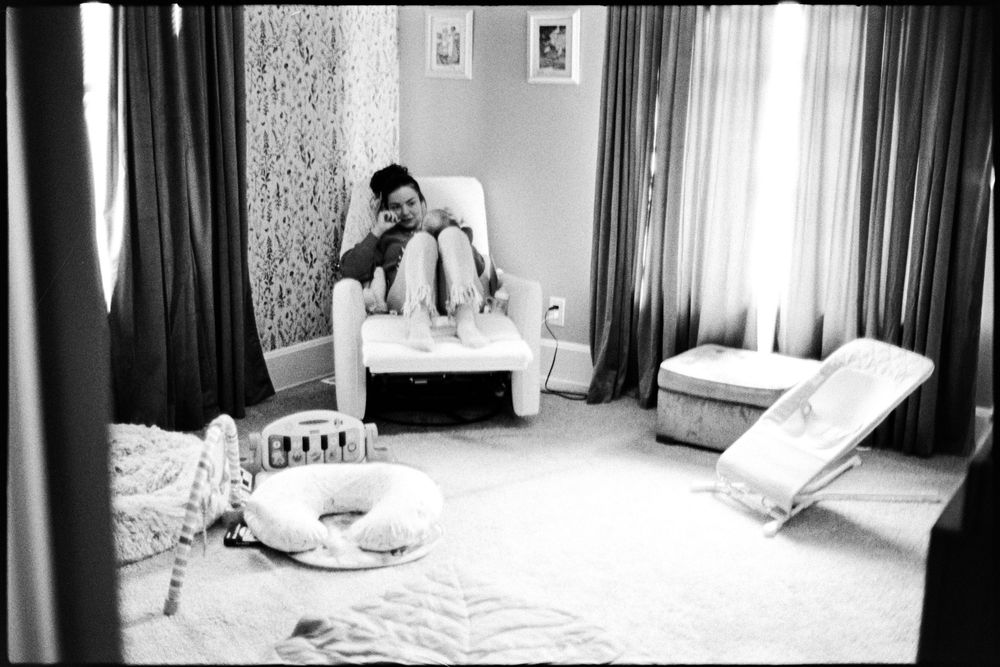 Person sits in nursery chair surrounded by baby items in black and white photo.