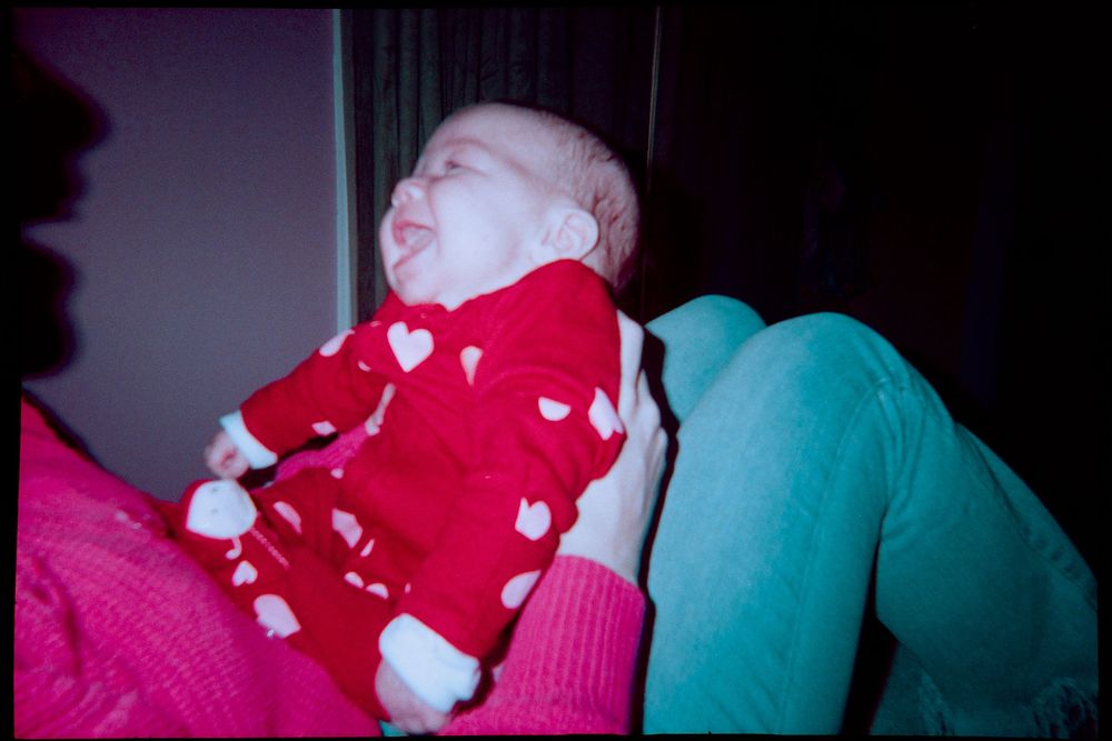 Newborn baby in red heart onesie resting against colorful cushions in soft home setting.