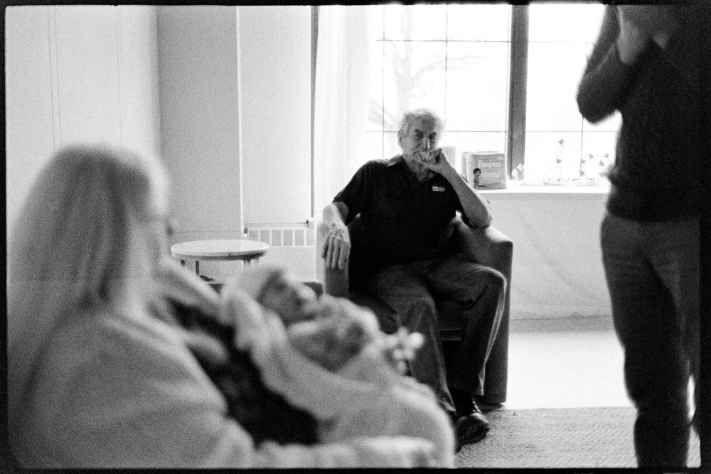 Elderly man seated by window in candid black and white family gathering photo with newborn.