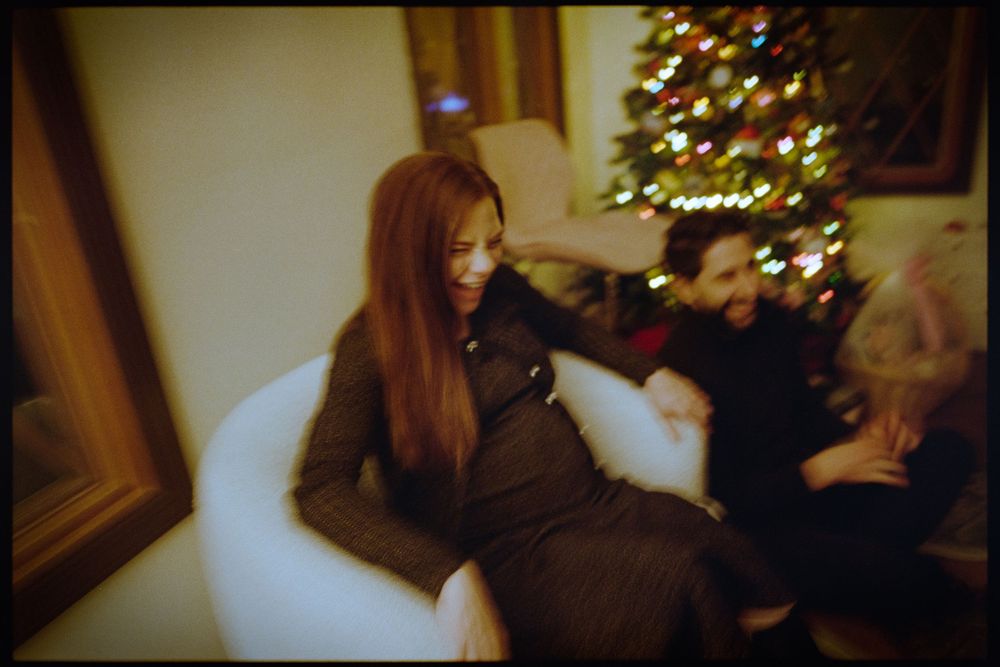 Woman with long red hair laughing joyfully while sitting in a white chair near a Christmas tree.