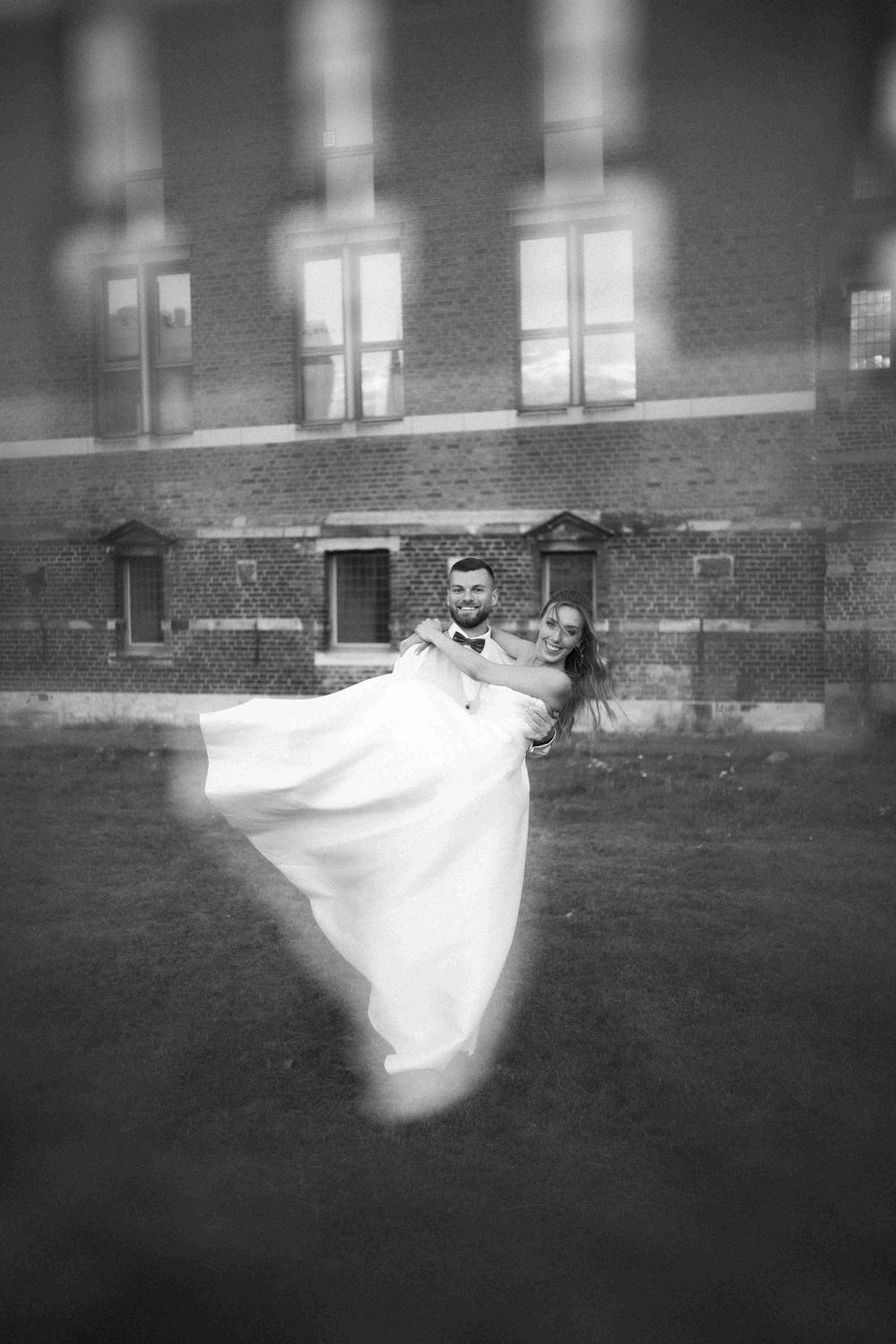 A blurred black and white image showing flowing white fabric movement across a grassy area near a brick building.