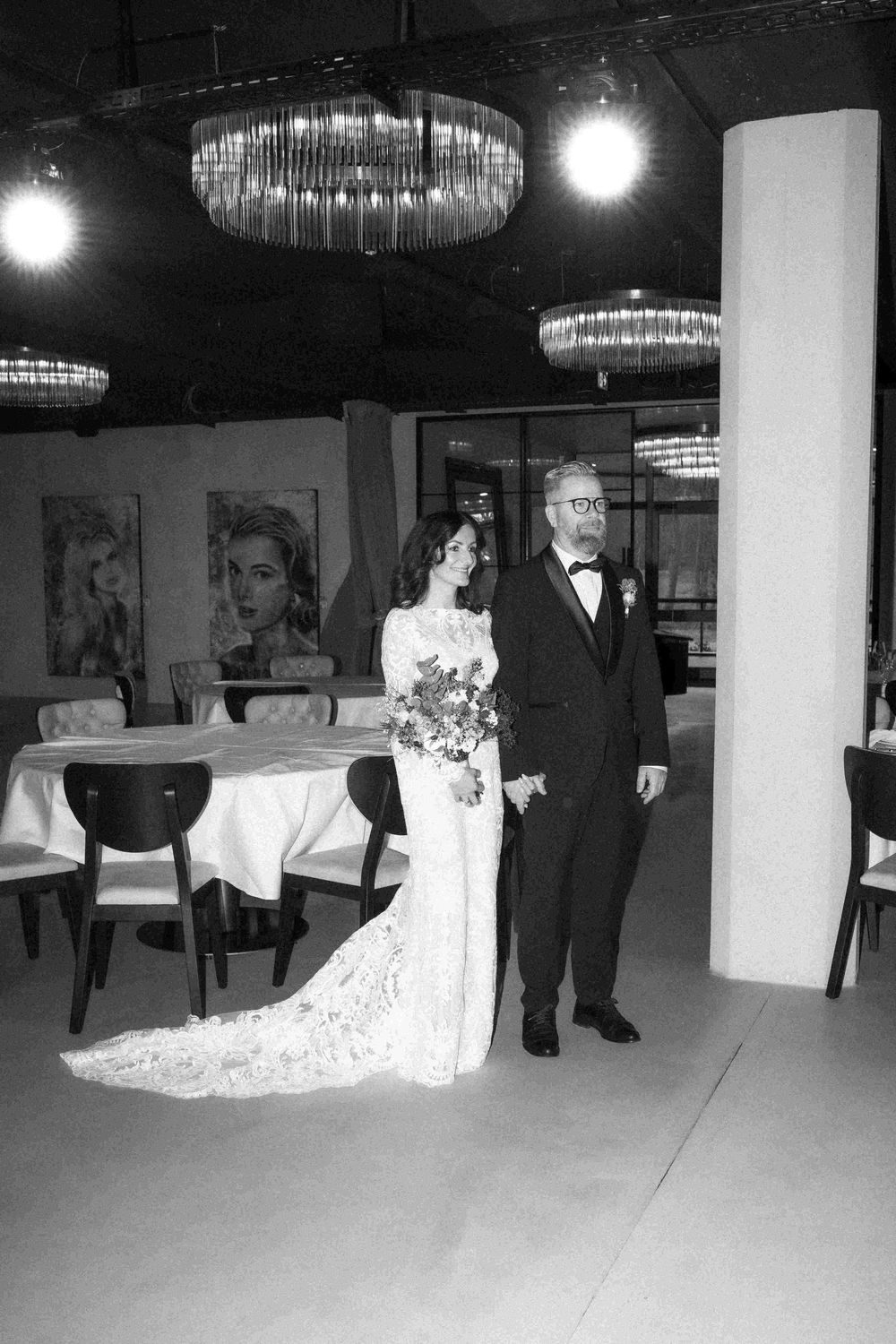 A couple stands in an elegant reception venue with crystal chandeliers and formal attire at their wedding celebration.