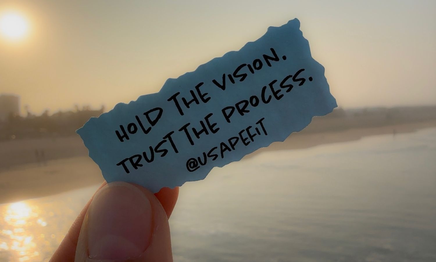 Hand holds up small blue note against sunset sky and ocean while sun creates reflective glow on calm water surface.