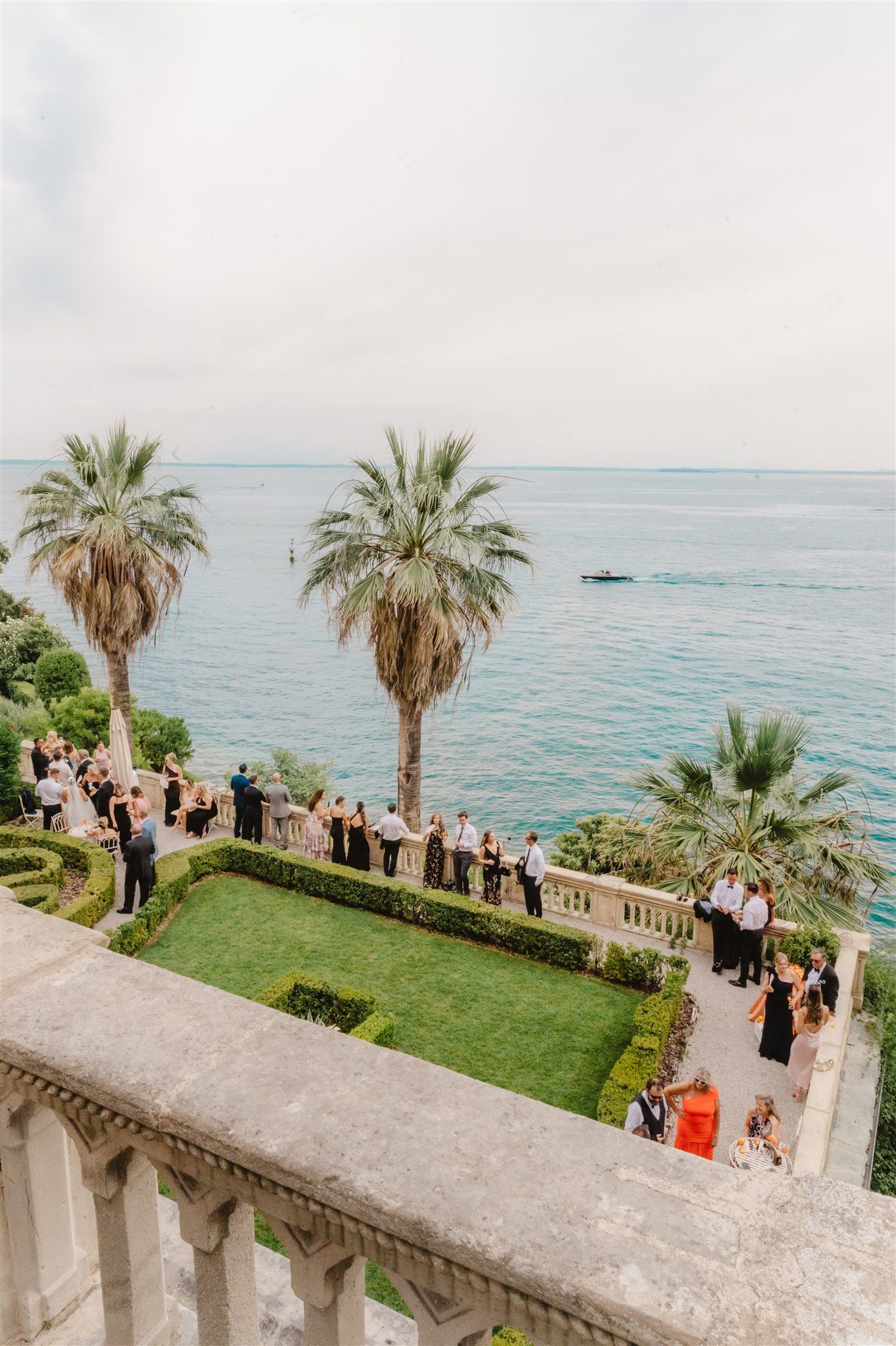 Wedding ceremony guests gathered on a Mediterranean terrace overlooking the sea with palm trees.