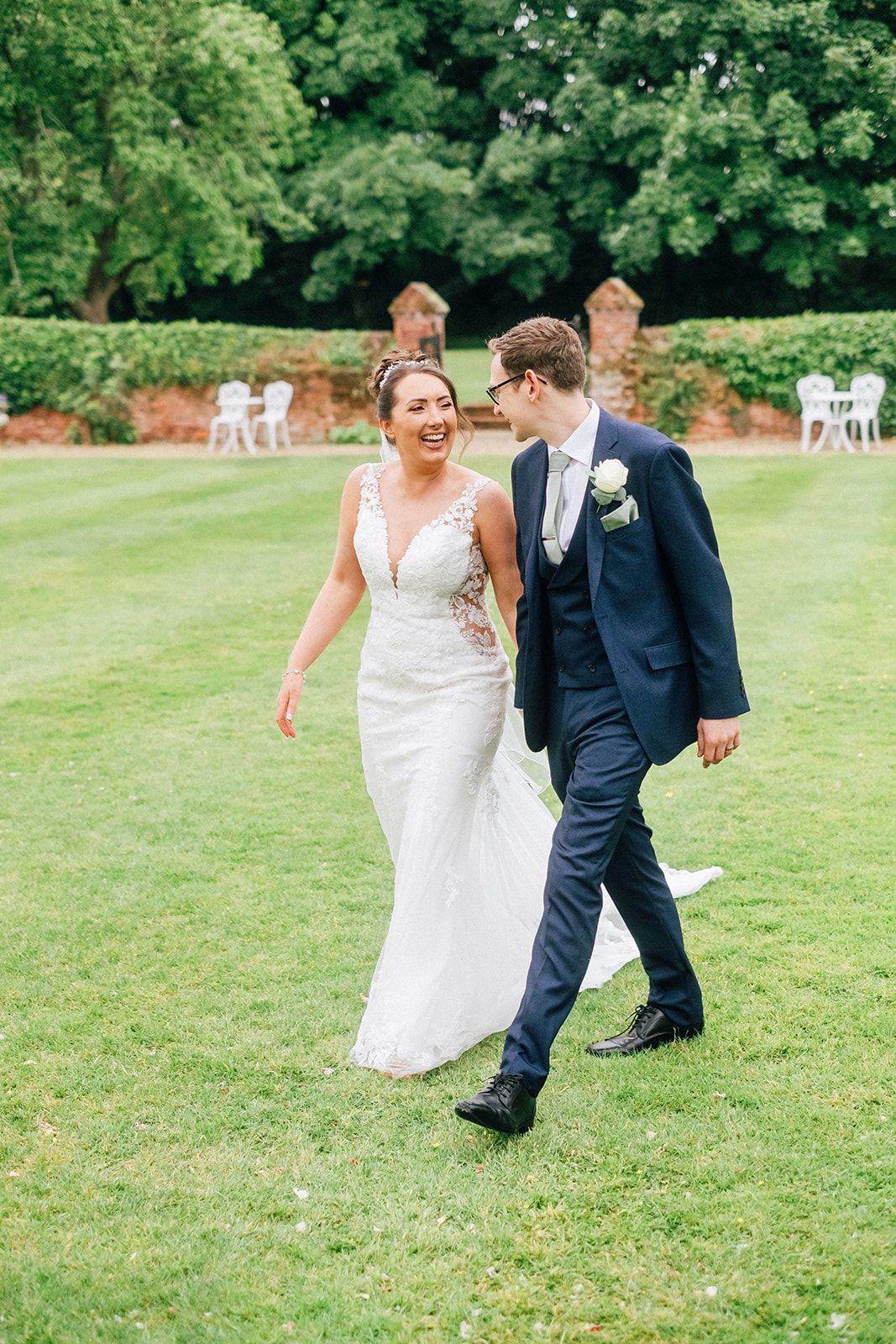 A joyful wedding couple walking and laughing together on a lush green lawn surrounded by trees.