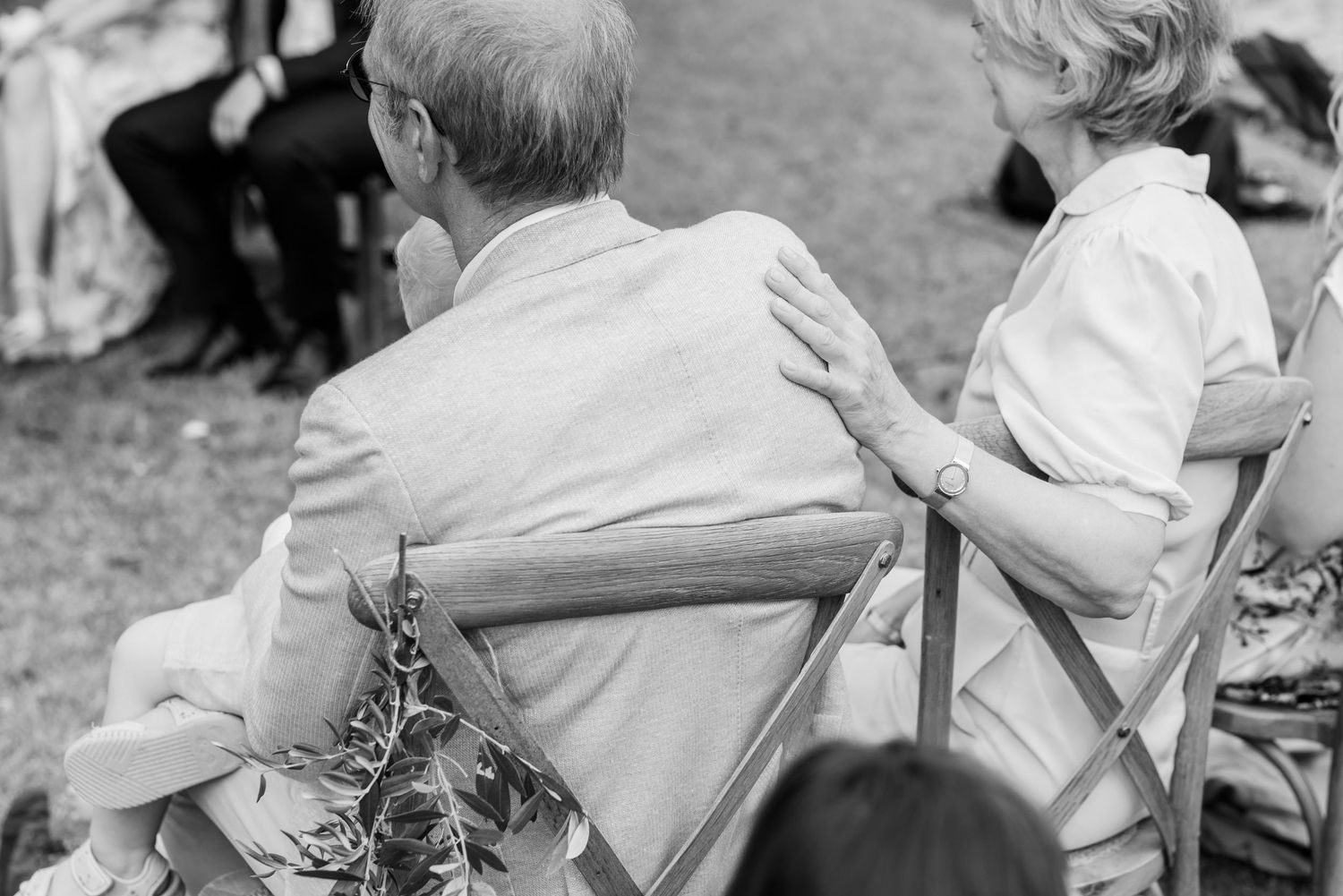 Schwarz-Weiß-Foto zeigt eine Dame, die ihre Hand zärtlich auf die Schulter ihres Mannes legt während sie der Trauung der Tochter lauschen.
