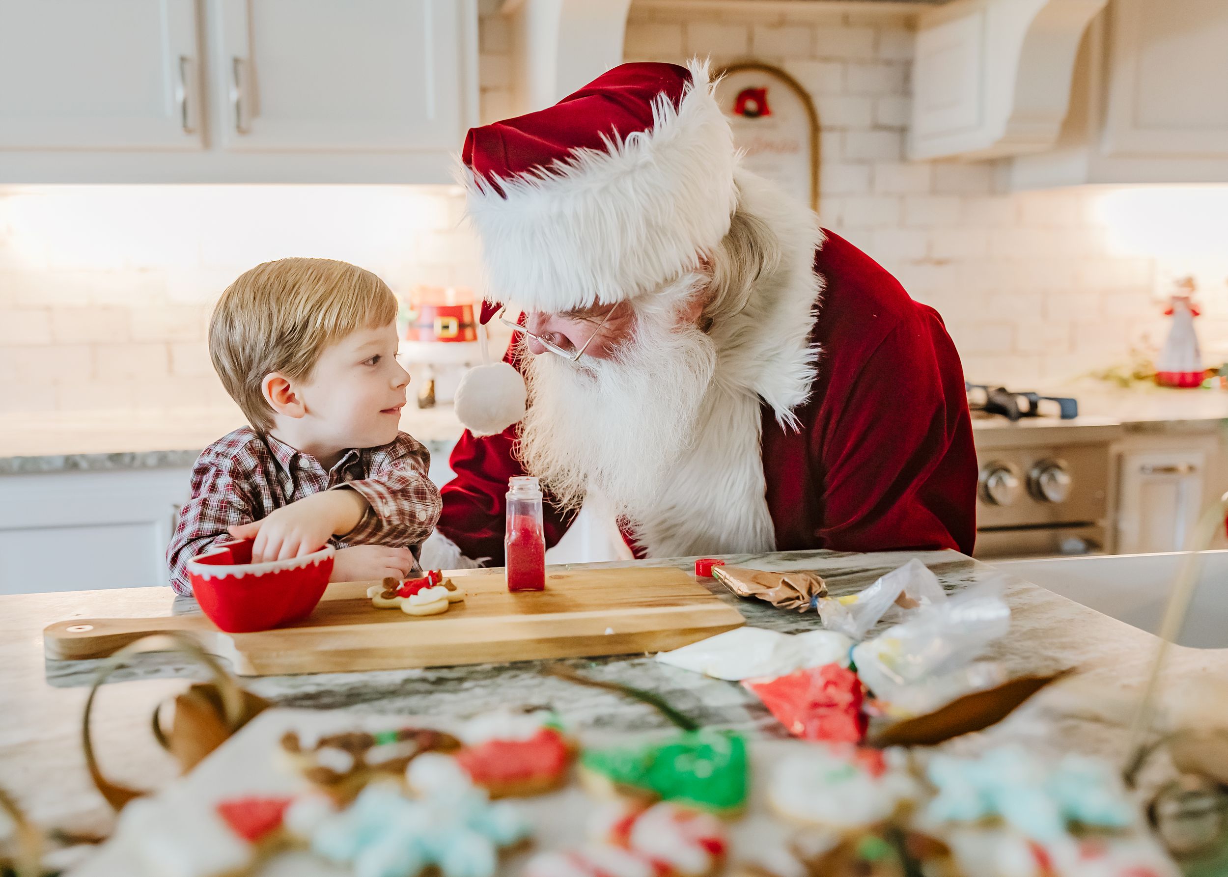 Holiday scene of a young child and Santa Claus baking cookies together in a bright kitchen with festive decorations.