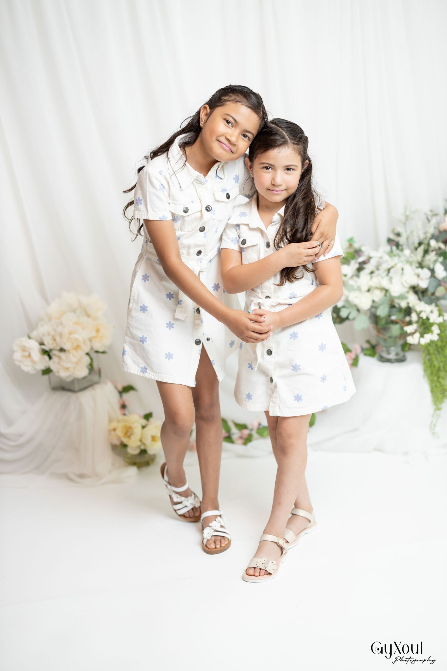 Two young children in matching white floral outfits pose together at a photo studio with floral decorations.