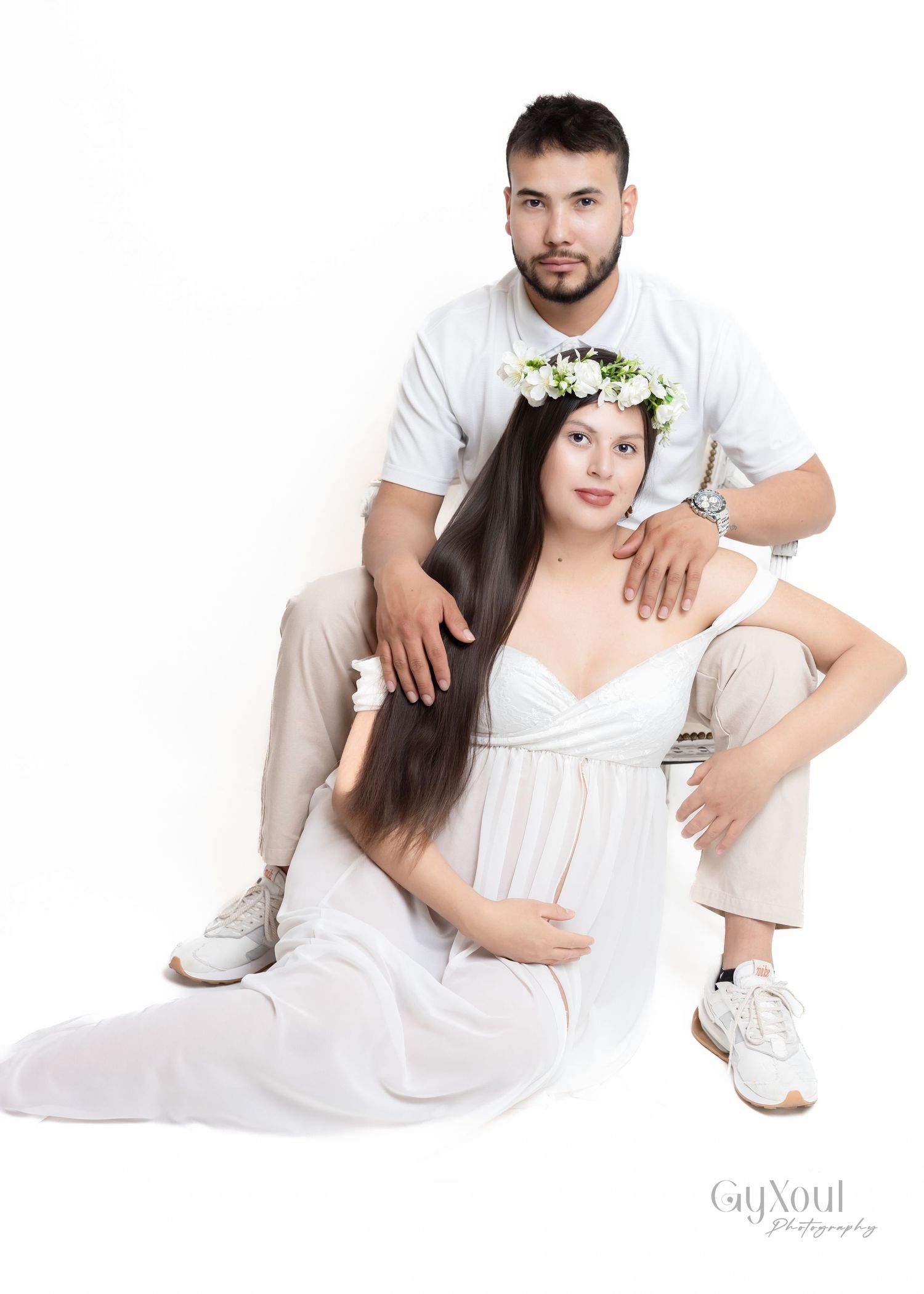 A couple poses together in a minimalist studio setting, wearing white outfits with the woman in a flowing dress and floral crown.