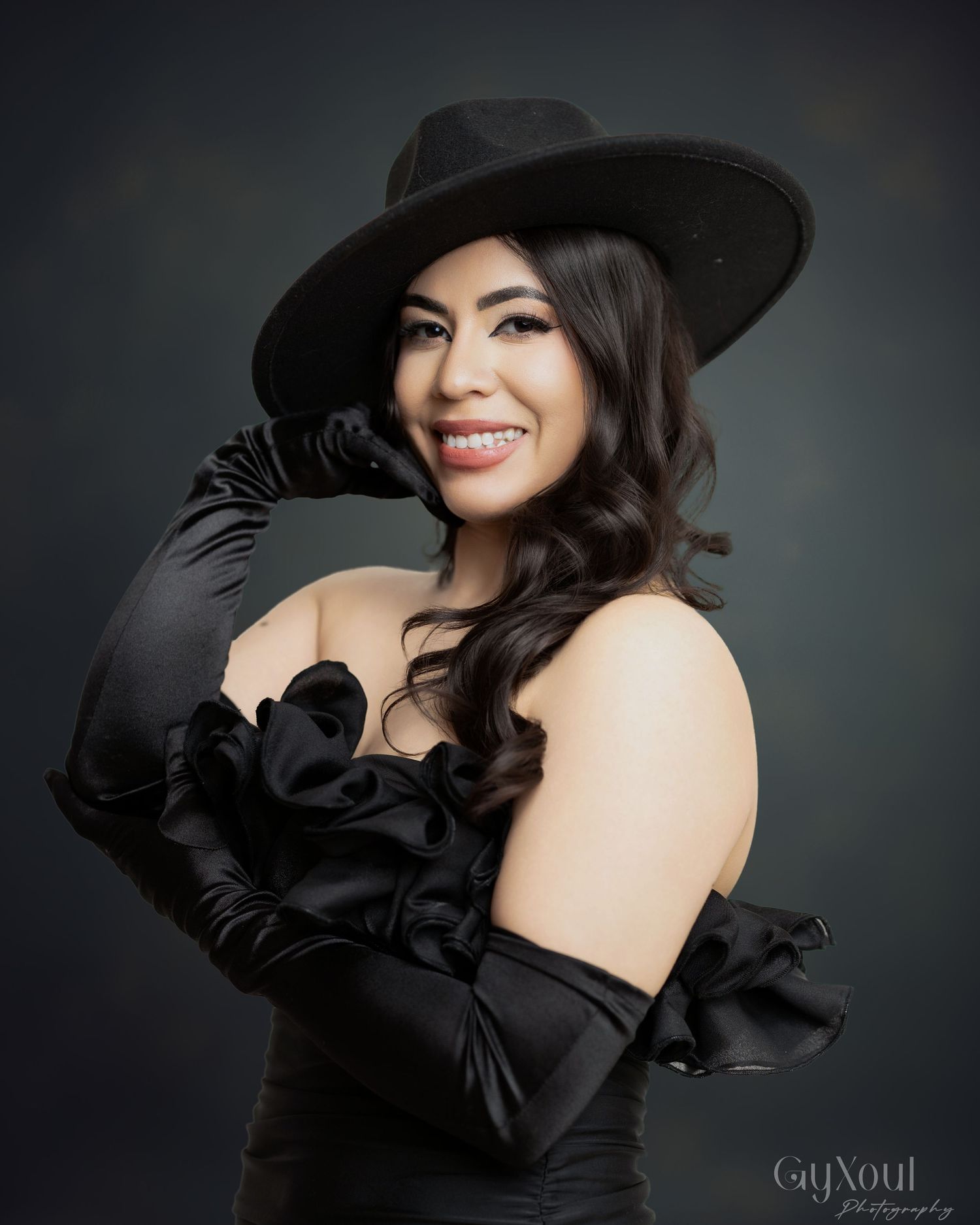 Studio portrait against dark backdrop featuring elegant black dress, wide brim hat, and dramatic backlit posing sequence.