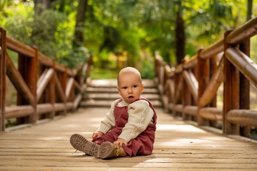 A baby lies on a wooden bridge in a lush green forest setting during a sunny day.