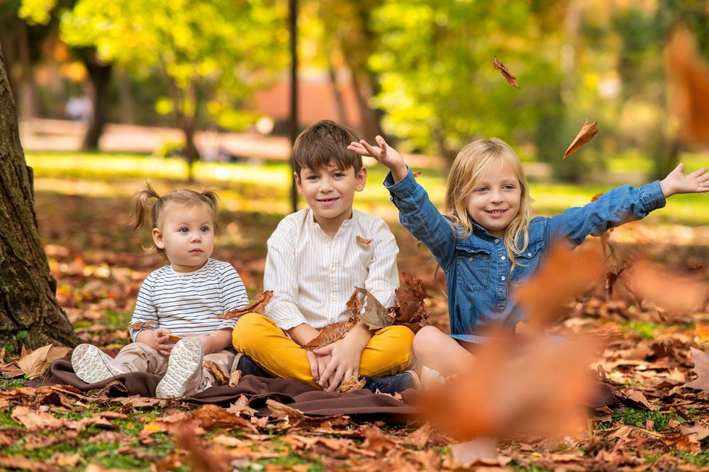 Three children sit among colorful fall leaves while playing and enjoying the autumn season outdoors.
