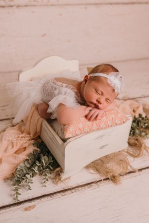 Sleeping baby in white lace outfit resting on vintage drawer with greenery decorations.