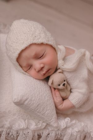 Sleeping baby in white knit bonnet cuddling small teddy bear on lace pillow.