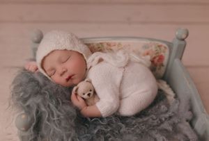 Sleeping infant in white outfit on vintage grey bed with textured blanket.