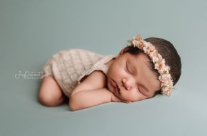 Sleeping infant wearing floral crown and knit wrap against grey background.
