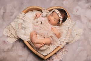 Newborn sleeping peacefully in heart-shaped basket wearing delicate lace dress and headband.