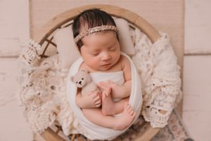 A sleeping newborn in a circular frame with lace wings and small teddy bear prop.
