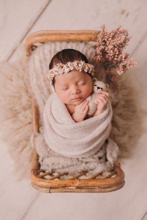 Sleeping newborn wrapped in beige blanket with floral crown and pink flowers on wooden chair.
