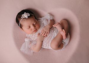 Sleeping newborn in delicate white lace dress with floral headband on pink background.