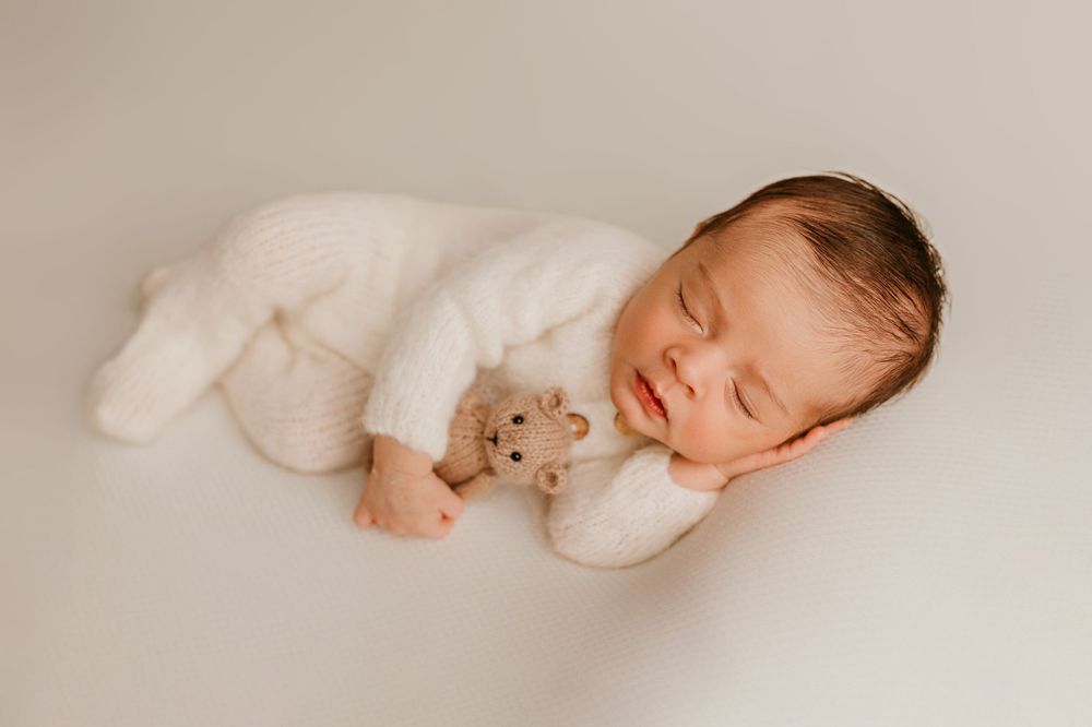 Sleeping newborn in white knit outfit cuddling small teddy bear on neutral background.