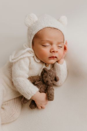 Peaceful sleeping baby in white knit outfit with bear ears holding brown teddy bear.