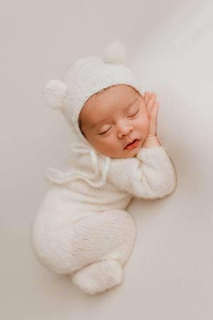 Sleeping newborn in white knit bear outfit with hand resting on cheek.