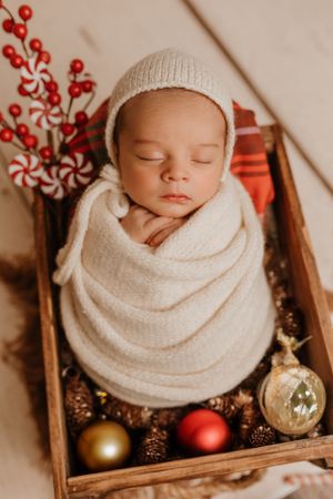Sleeping newborn wrapped in white knit blanket in rustic wooden box with Christmas ornaments and red berries.