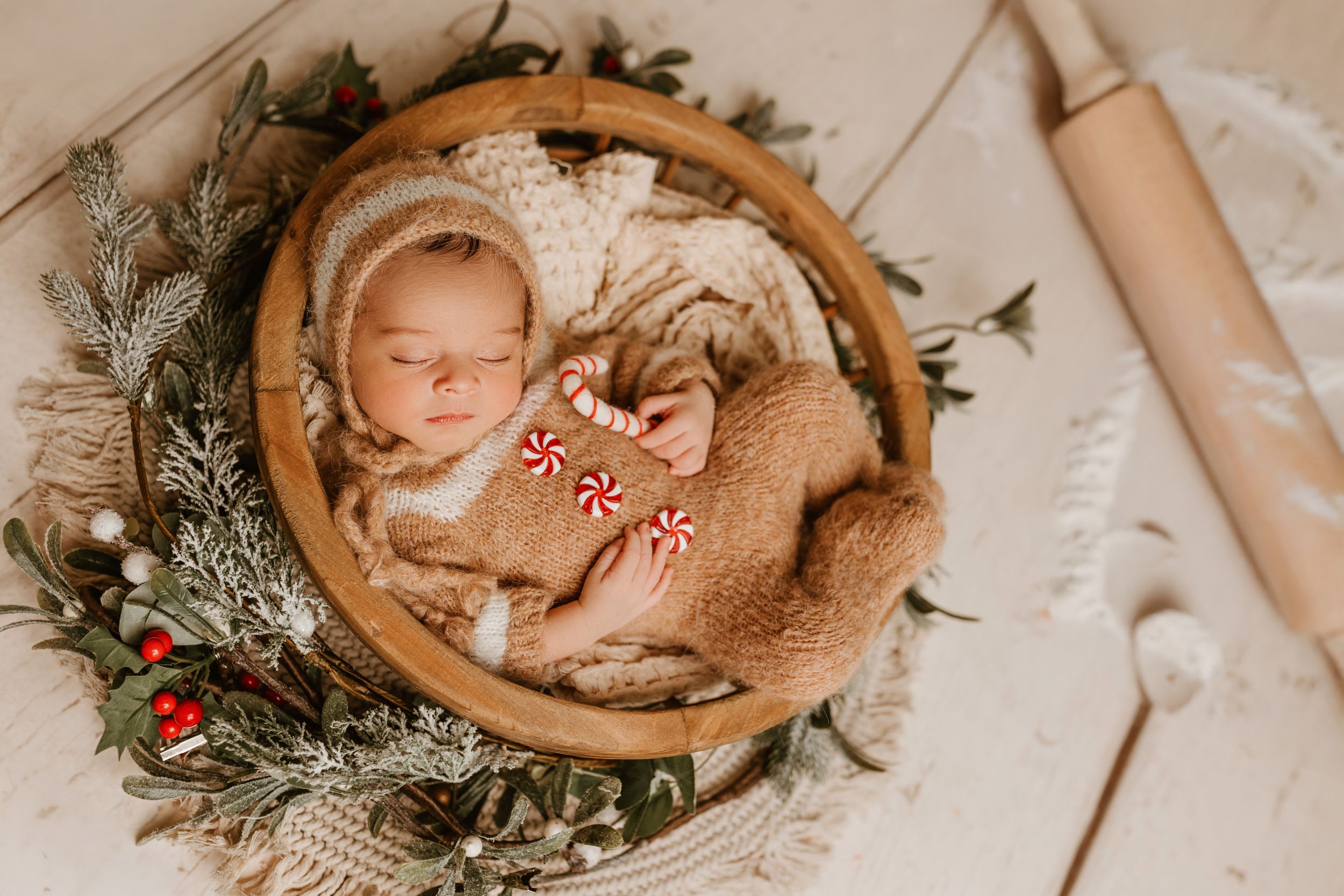 Sleeping newborn wrapped in blanket in wooden bowl surrounded by winter greenery and berries.
