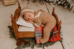 Sleeping newborn in cozy brown outfit on miniature wooden bed with plaid blanket and holiday decor.