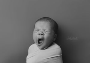 Newborn in white wrap yawning against gray backdrop in black and white portrait.