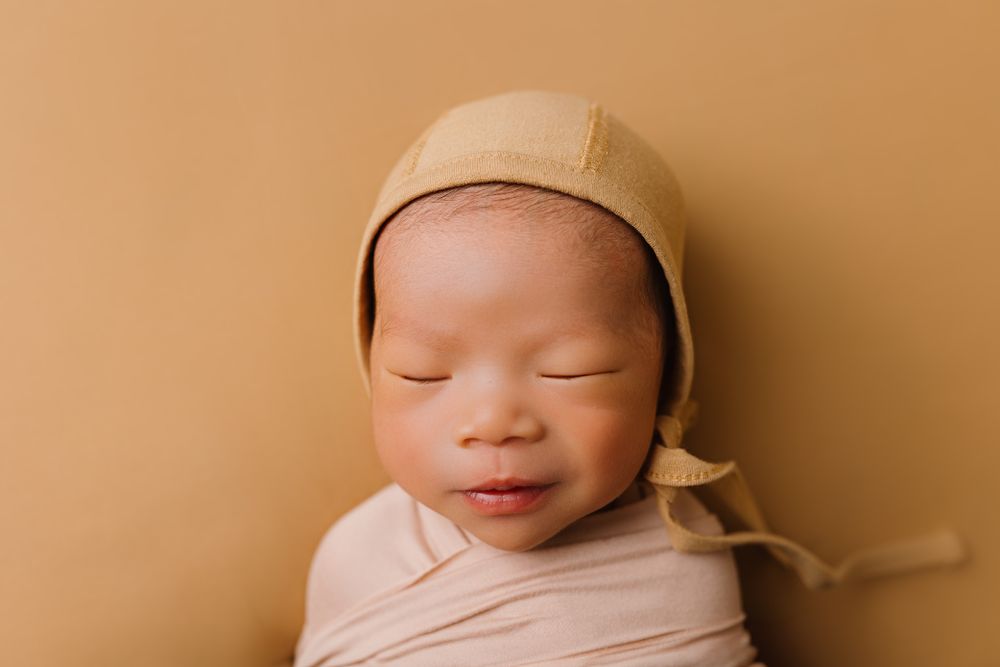 Sleeping baby wrapped in neutral toned swaddle with beige bonnet against warm background.