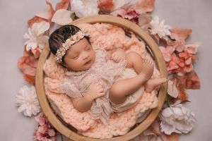 Newborn sleeping peacefully in a wooden bowl surrounded by pink and white floral decorations.