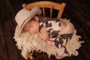 Sleeping newborn in cowboy hat and western outfit on wooden chair with textured blanket.