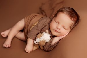 Sleeping infant in brown outfit with small plush toy on warm brown background.